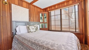 Une chambre avec un plafond en bois, un lit avec une literie à motifs, une table de chevet, une armoire avec des portes en verre et une fenêtre avec des stores.