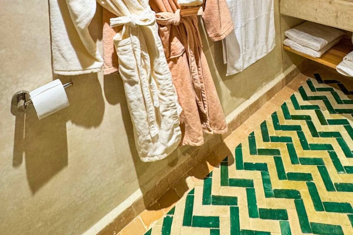 A bathroom with a towel rack holding two robes, a toilet paper roll, and a mirror above a sink with a tiled floor.