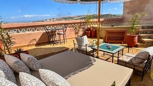 Une terrasse avec une table, des chaises et un grand parasol. On y trouve des plantes en pot et une vue sur une ville lointaine.