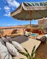 Terraço com mesa, cadeiras e um grande guarda-sol. Há plantas em vasos e vista para uma cidade ao longe.