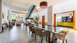 Amplia sala de estar con una mesa de comedor de vidrio, asientos cómodos, una escalera moderna y una cocina luminosa al fondo.