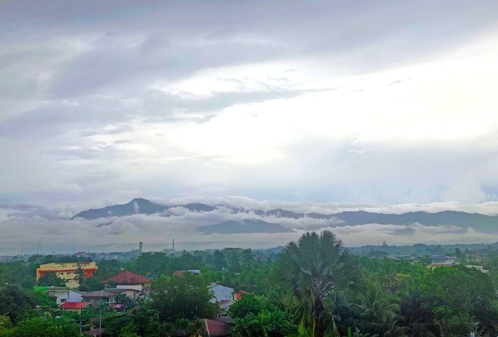 Eine Landschaft mit einem Gebirge, Wolken und einer Stadt mit Gebäuden und Grünflächen.
