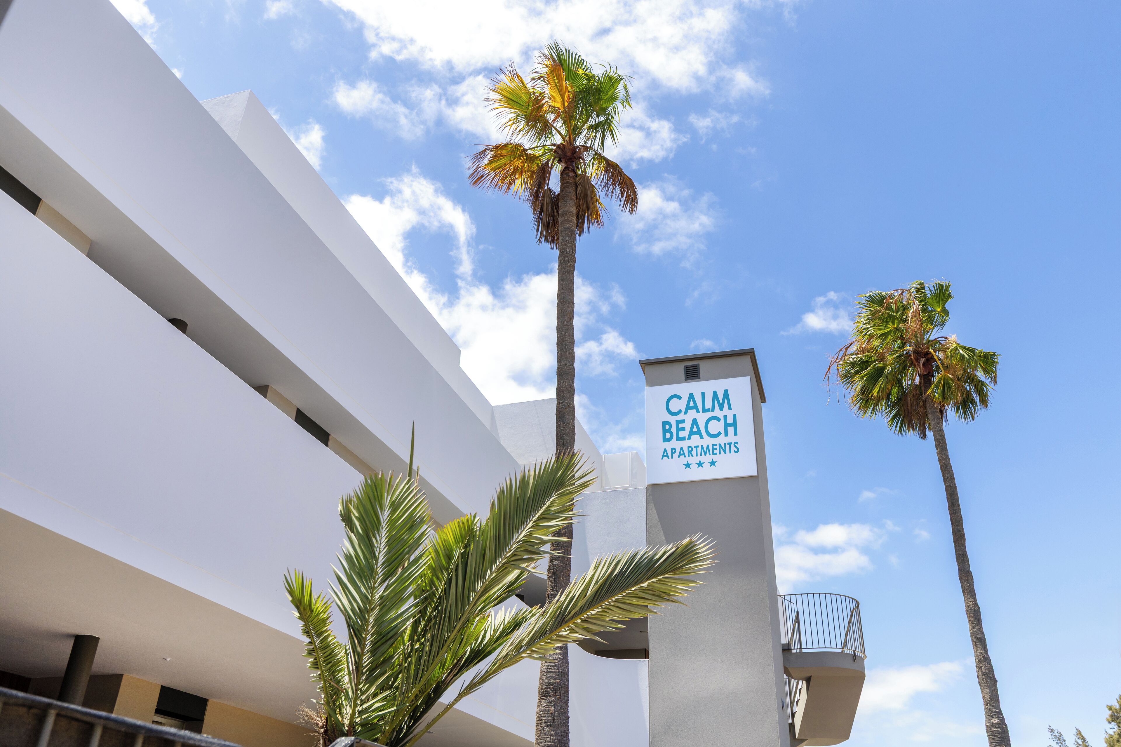 Een modern gebouw met een bord waarop 'CALM BEACH APARTMENTS' staat en palmbomen ervoor.