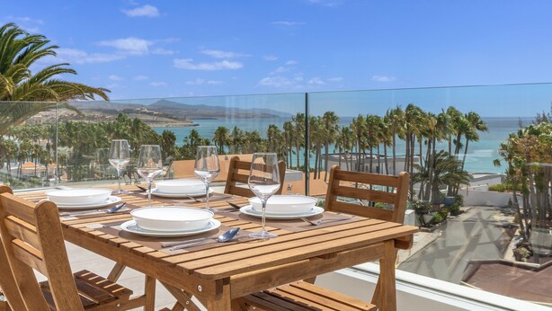 Una mesa de madera lista para comer, con vista al océano y palmeras.