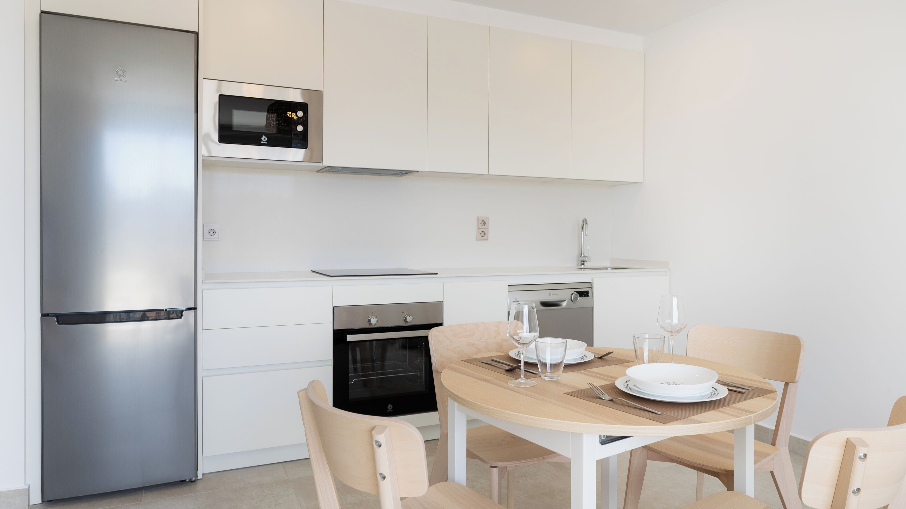 A modern kitchen with a stainless steel refrigerator, a microwave, a built-in oven, and a dishwasher. There is a wooden dining table with chairs, and a glass of water and a wine glass on the table.