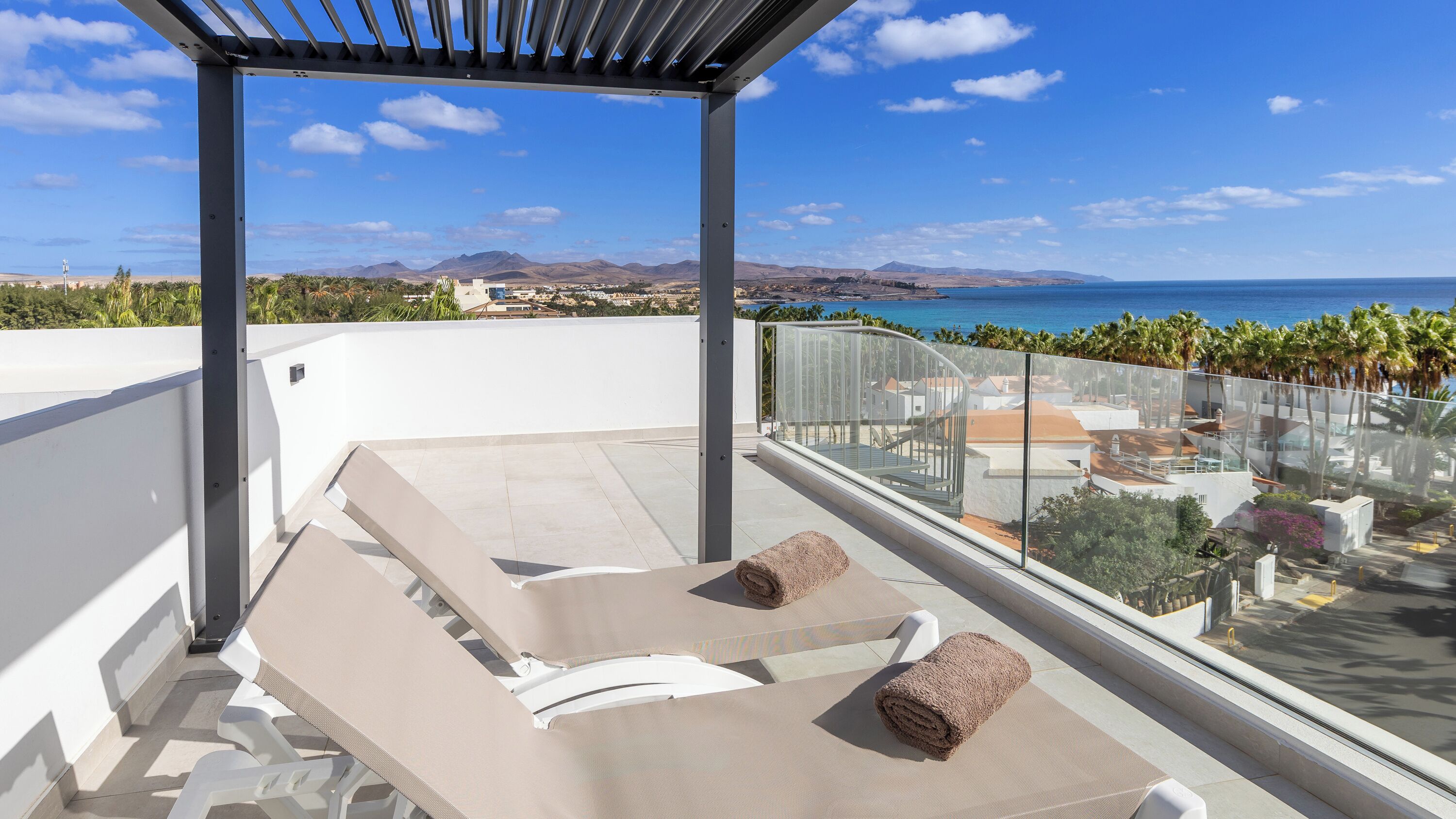 A rooftop terrace with two sun loungers, a pergola, and a view of the ocean and mountains.