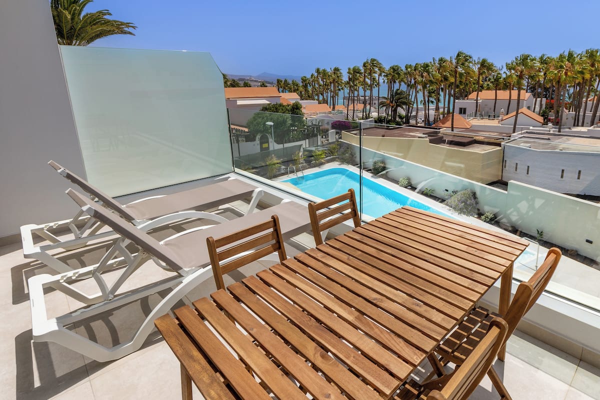 A balcony with a wooden table and chairs, a glass railing, and a view of a swimming pool and palm trees.