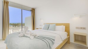 Una habitación de hotel con una cama grande, vistas a la playa y una mesita de noche de madera.