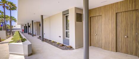 A modern building corridor with a row of doors, a planter with plants, and a clear sky.