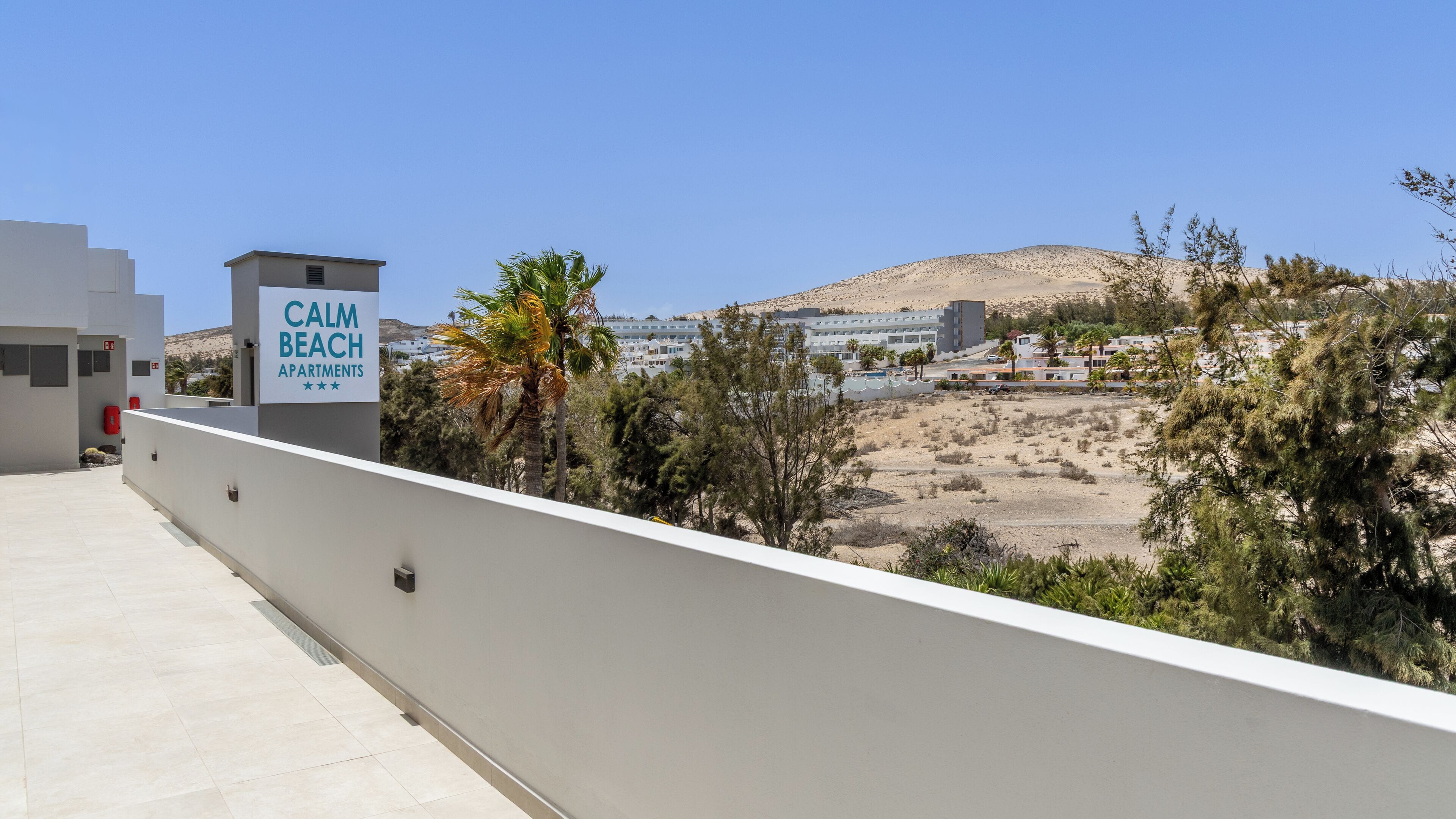 A sign for 'Calm Beach Apartments' with a view of a desert landscape and buildings in the background.