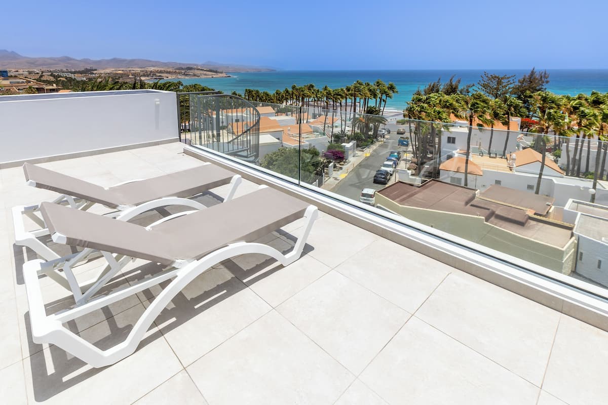 A rooftop terrace with two lounge chairs and a glass balustrade overlooking a coastal area with palm trees and buildings.