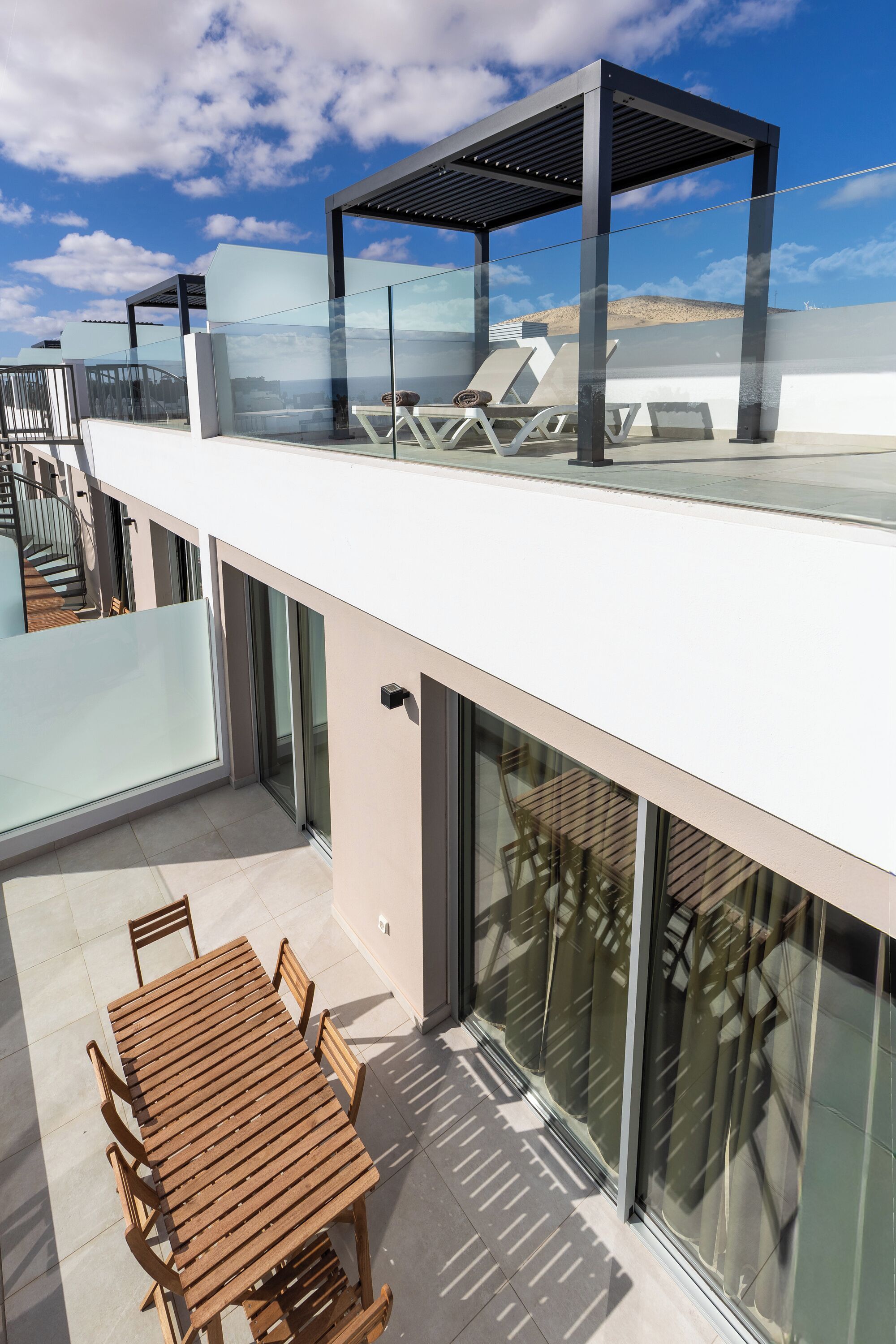 Une terrasse extérieure moderne dotée d’une balustrade en verre, d’une table en bois avec des chaises, et d’un espace détente avec des fauteuils.