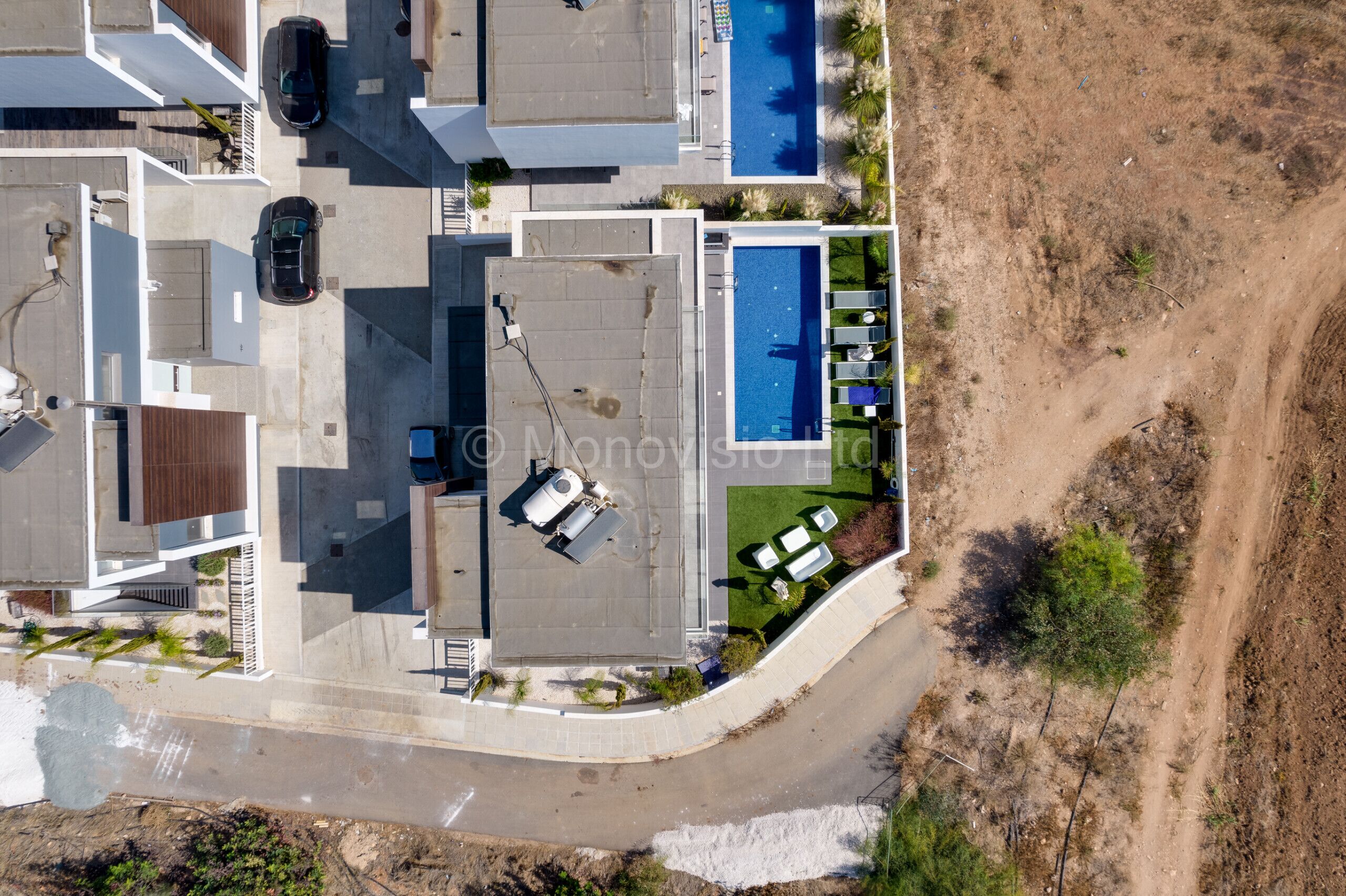 Vista aérea de uma área residencial moderna com piscina, vários carros e uma estrada de terra.