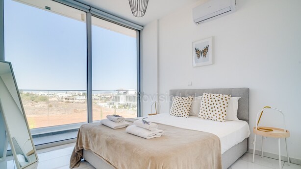 Une chambre moderne avec une grande fenêtre, un lit recouvert d’une couverture beige et une petite chaise en bois.