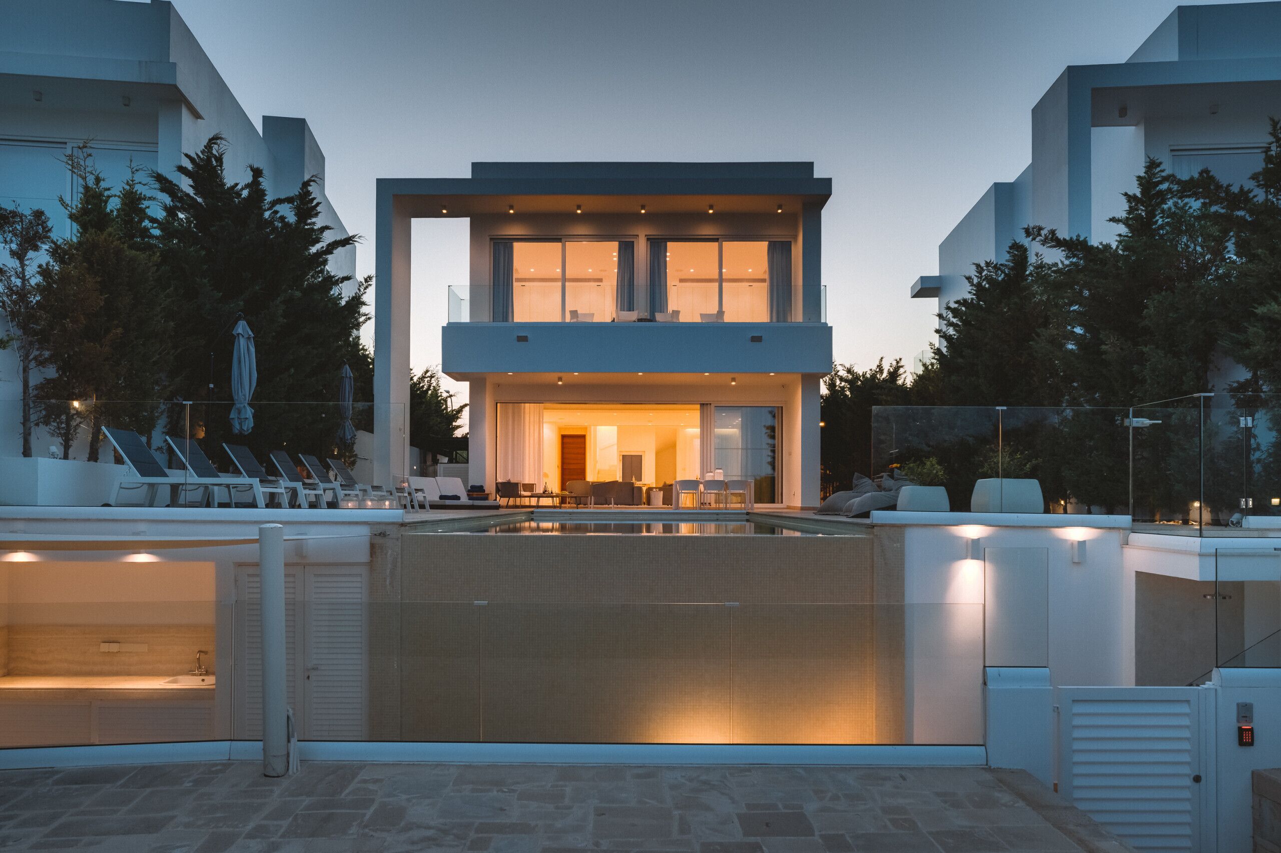 A modern two-story building with a swimming pool, lounge chairs, and a well-lit entrance.