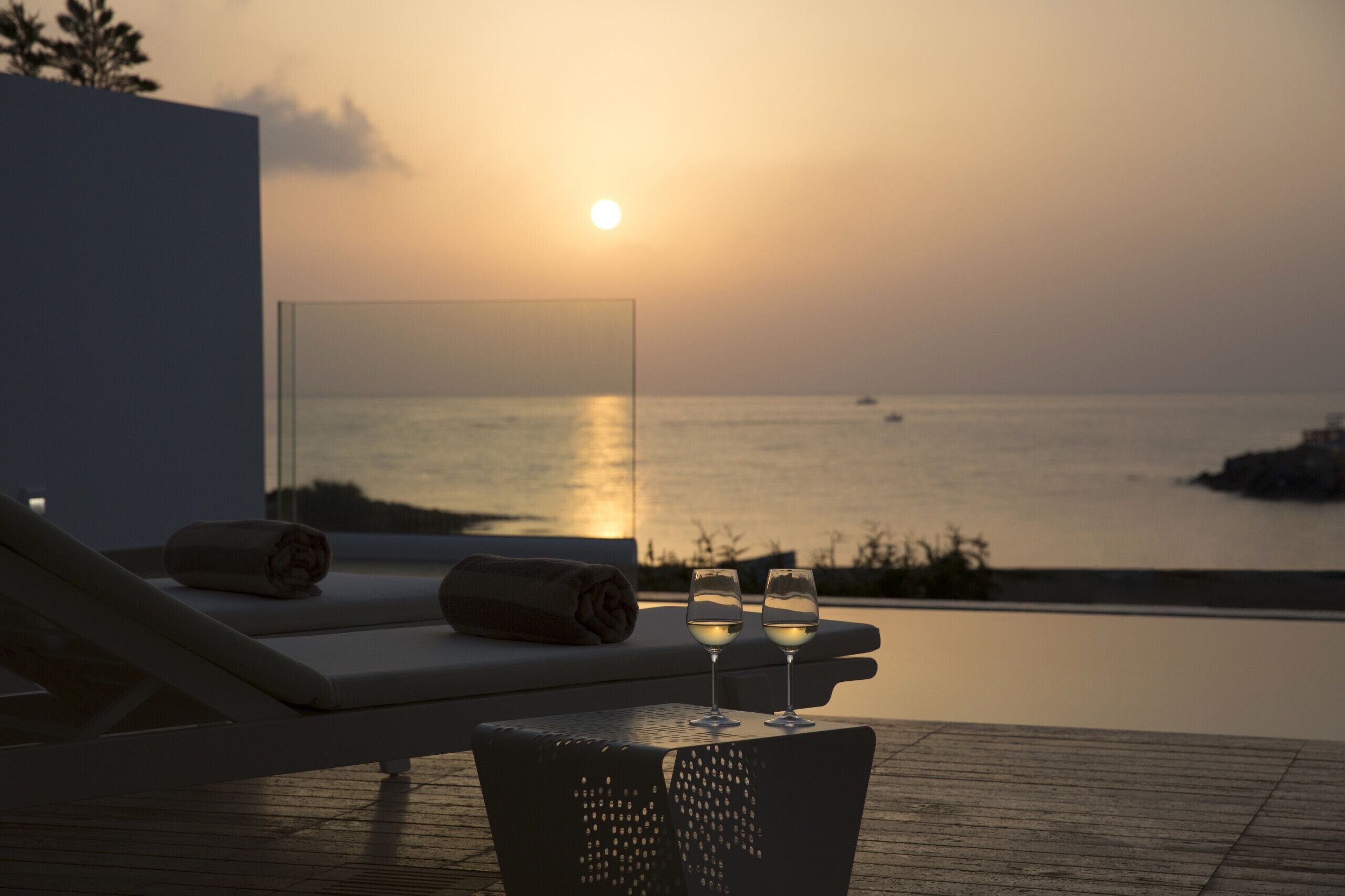 Vista del atardecer desde una zona de piscina con sillones de descanso y dos copas de vino blanco.
