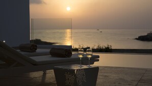Vista do pôr do sol de uma área com piscina, cadeiras de espreguiçadeira e dois copos de vinho branco.