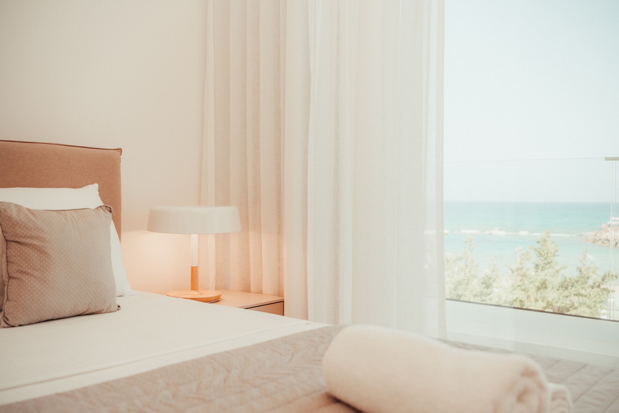 A hotel room with a bed, a nightstand with a lamp, and a view of the sea.
