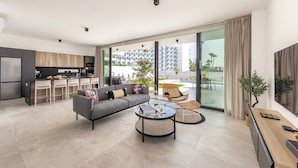 Una sala moderna con sofá, mesa de centro y sillas, un área de cocina con comedor, y grandes puertas corredizas de vidrio que dan a un balcón con vista a la piscina.