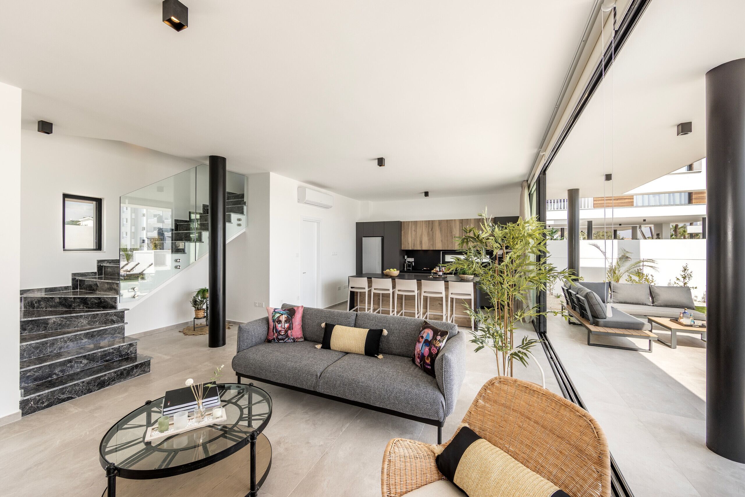 Un salon moderne avec un canapé gris, une table basse en verre et un fauteuil en osier. Un escalier mène à un espace cuisine équipé d’un coin repas.