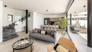 Una sala de estar moderna con un sofá gris, una mesa de centro de cristal y una silla de mimbre. Una escalera conduce a una zona de cocina con un comedor.