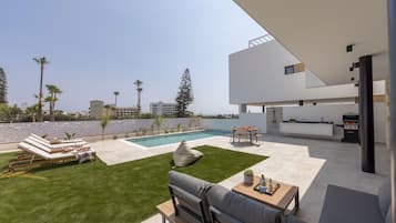 Un espacio exterior moderno con piscina, sillones de descanso, una zona de barbacoa y un área de cocina.