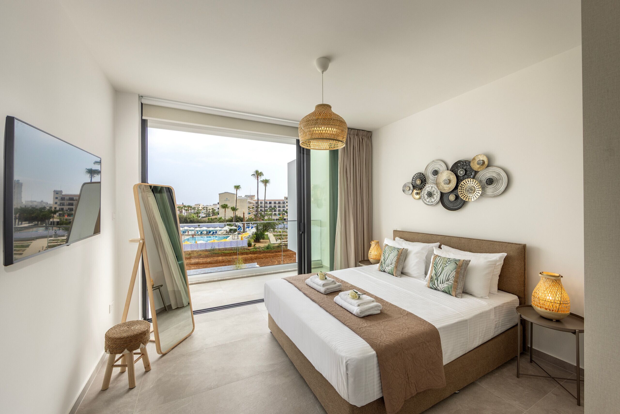 A modern hotel room with a large bed, a painting easel, and a view of a pool and buildings.