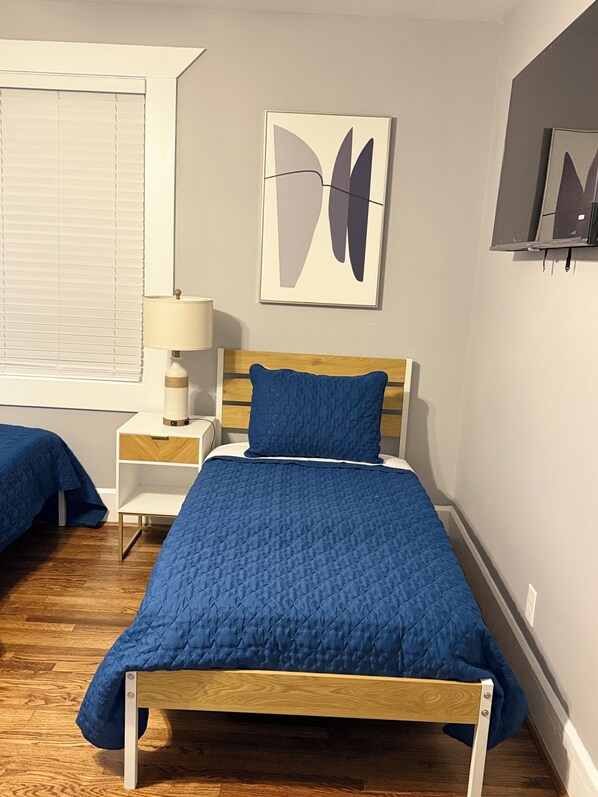 A bedroom with a wooden bed, a nightstand with a lamp, a window with blinds, and a wall-mounted shelf with a framed picture. - Catherine’s Castle – Your Home Away from Home! (Birmingham)