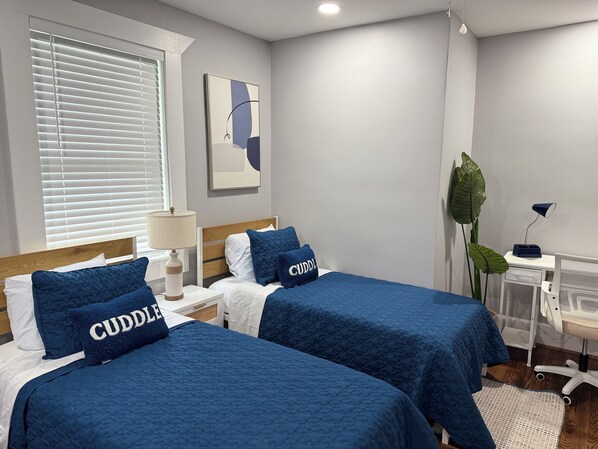 A hotel room with two beds, each with a blue quilted bedspread and pillows labeled "CUDDLE". There is a bedside table with a lamp, a large green plant, and a desk with a chair.