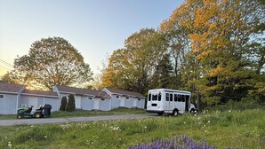 Una caravana blanca estacionada en un césped con flores moradas al frente, una hilera de casas móviles en el fondo y árboles con hojas otoñales.
