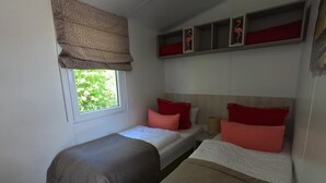 A small, compact room with two single beds, a window with a patterned valance, and a shelf with decorative items. - Shell on the Rhön beach with whirlpool (Wartmannsroth)