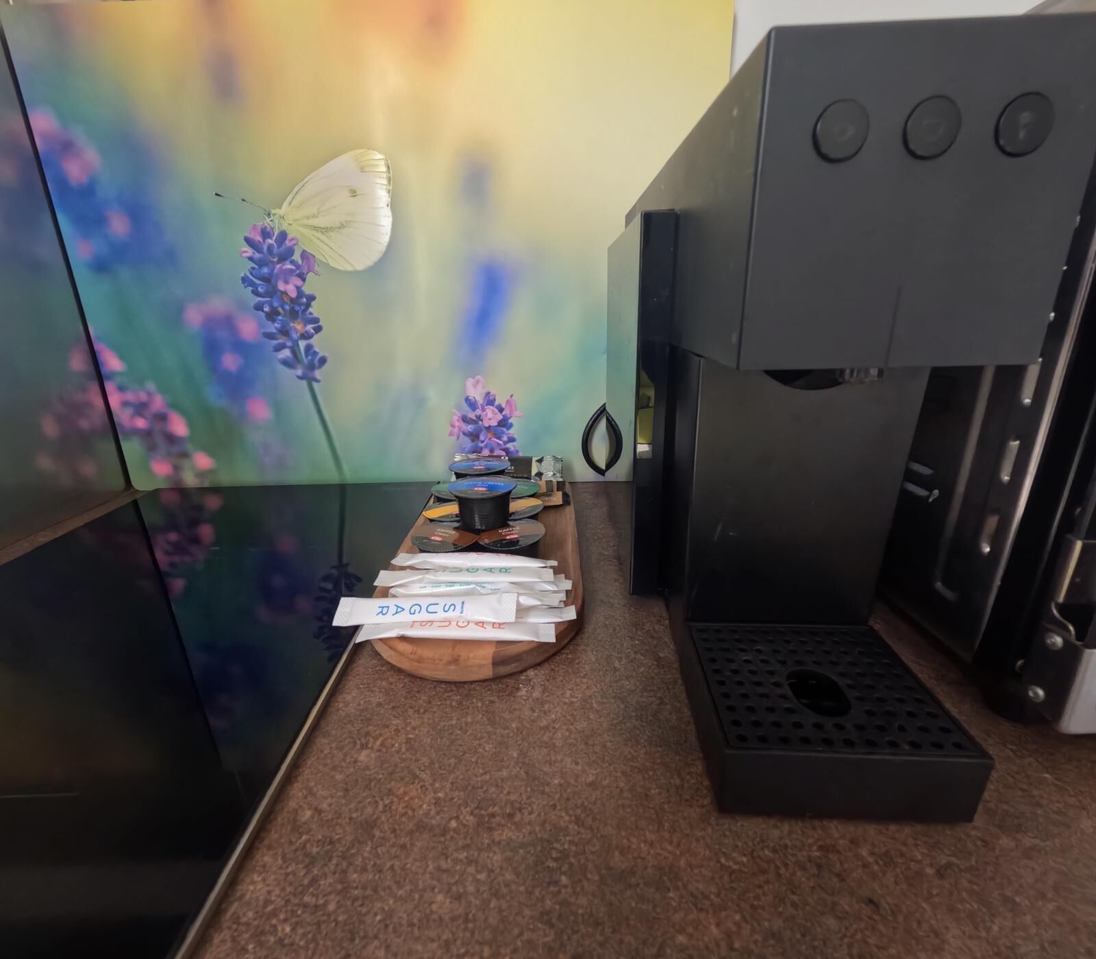 A coffee machine, a wooden tray with coffee pods, and a wall with a floral design.