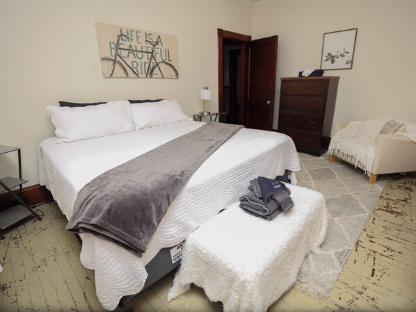 Una cama bien hecha con un edredón gris, un tapete blanco y mullido, y un pequeño banco con toallas dobladas.