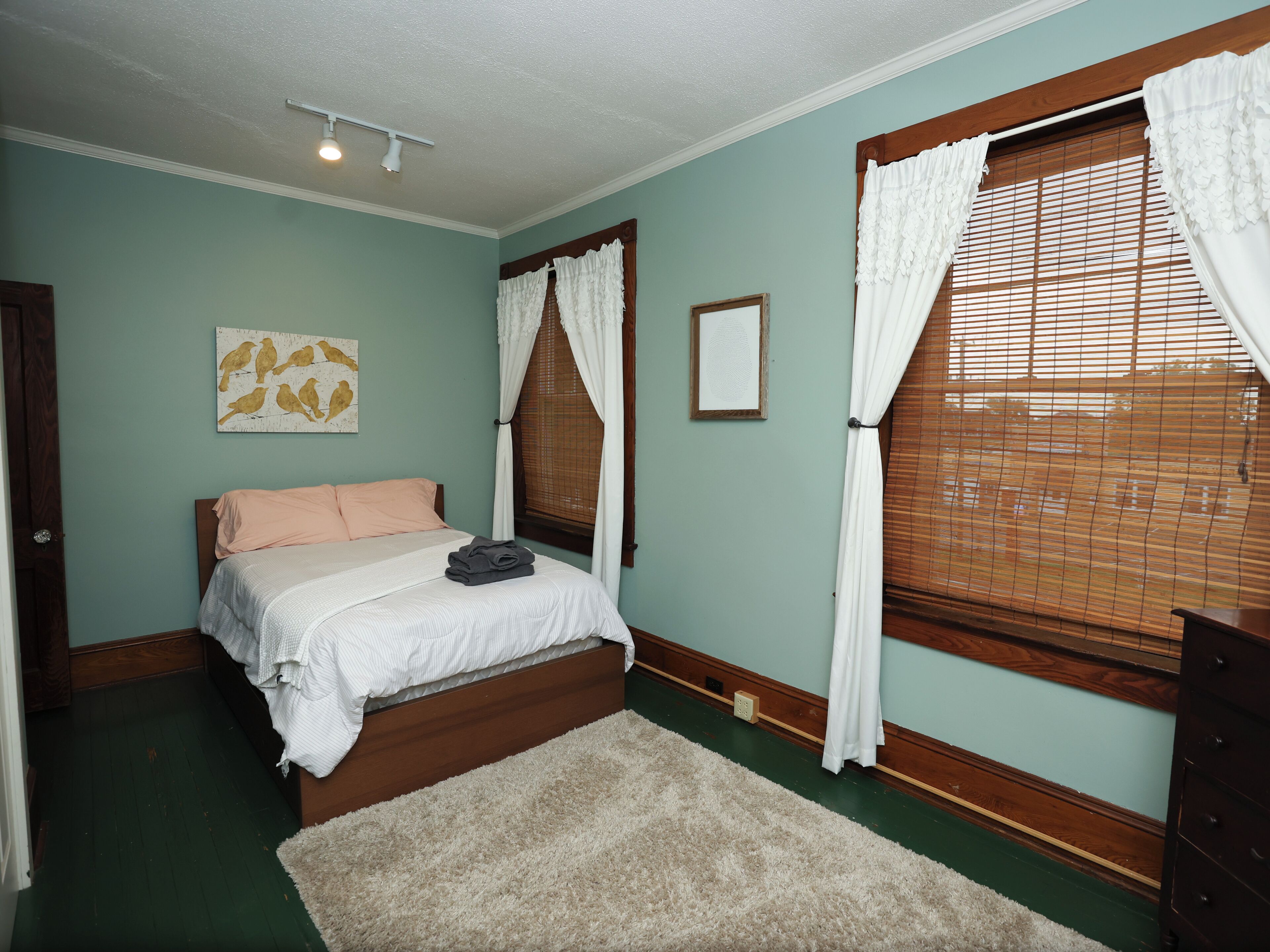 A bedroom with a bed, a nightstand, a dresser, a window with blinds, and a wall-mounted light fixture.