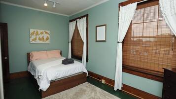 A bedroom with a bed, a nightstand, a dresser, a window with blinds, and a wall-mounted light fixture.