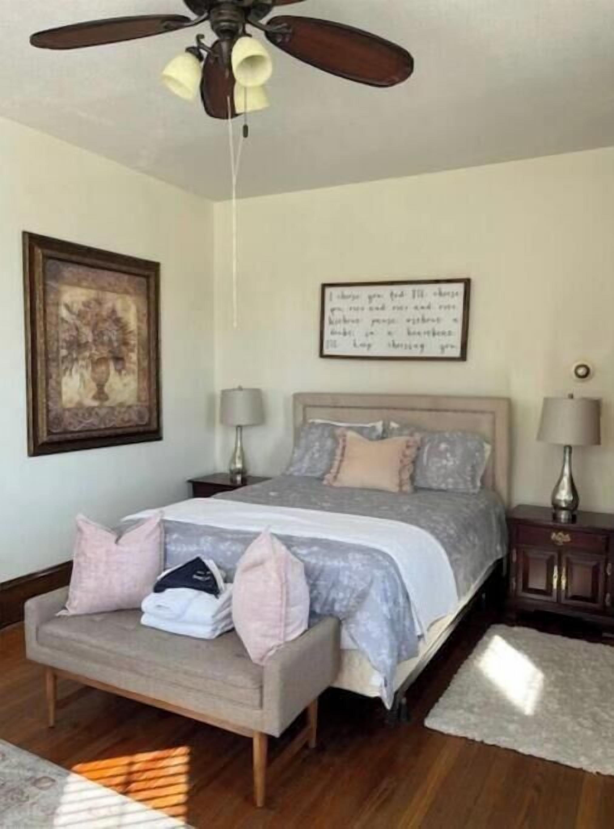 Una camera da letto con un letto, un comodino, una lampada, una ventola a soffitto, un quadro incorniciato e una frase motivazionale sulla parete.