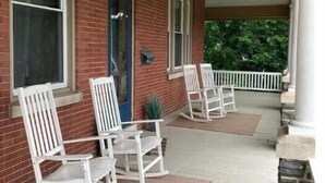 A covered porch with white rocking chairs, a blue door, and a brick wall. - Historic Main Street Getaway (Leola)