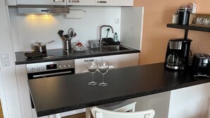 Ett kompakt kök med vita skåp, en svart bänkskiva och ett trägolv. Det finns två vita stolar, en kaffebryggare och en diskho med blandare.