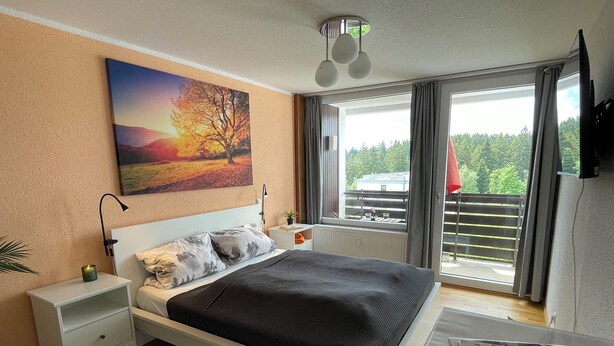 Ett sovrum med en säng, sängbord, ett litet bord, ett fönster med gardiner, en taklampa och en väggmonterad TV.