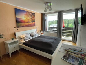 Une chambre à coucher comprenant un lit, des tables de chevet, une commode, une fenêtre avec des rideaux, un luminaire au plafond et un téléviseur fixé au mur.