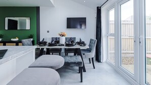 A modern dining area with a table set for four, a television mounted on the wall, and a view of the backyard through large windows.