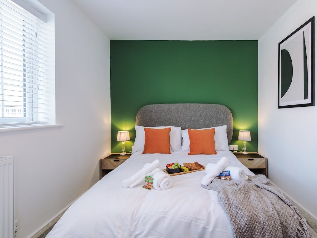 A neatly made bed with white linens, orange pillows, and a green headboard. There is a tray with breakfast items and towels on the bed.