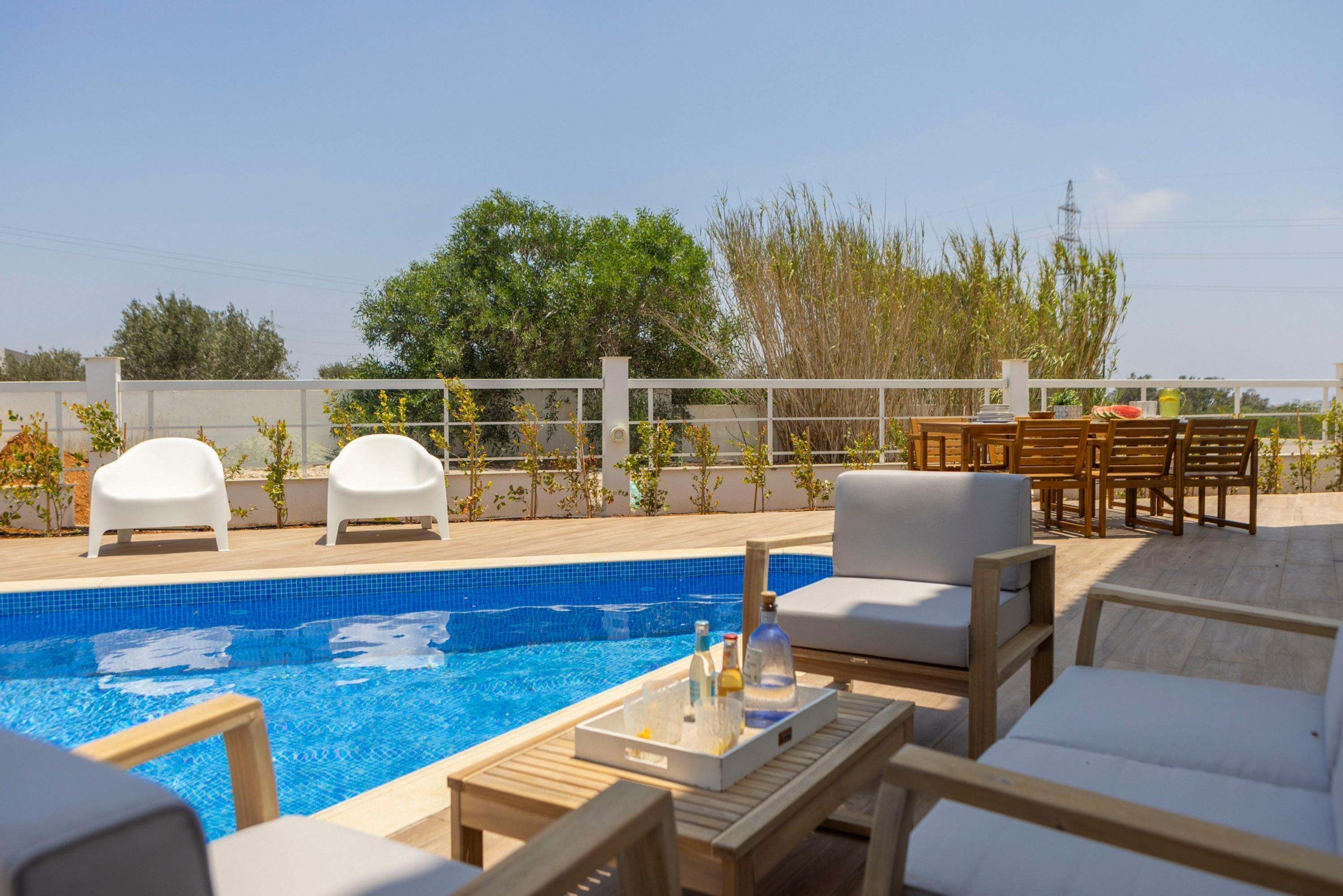 A rooftop pool area with lounge chairs and tables, surrounded by a white fence and vegetation.