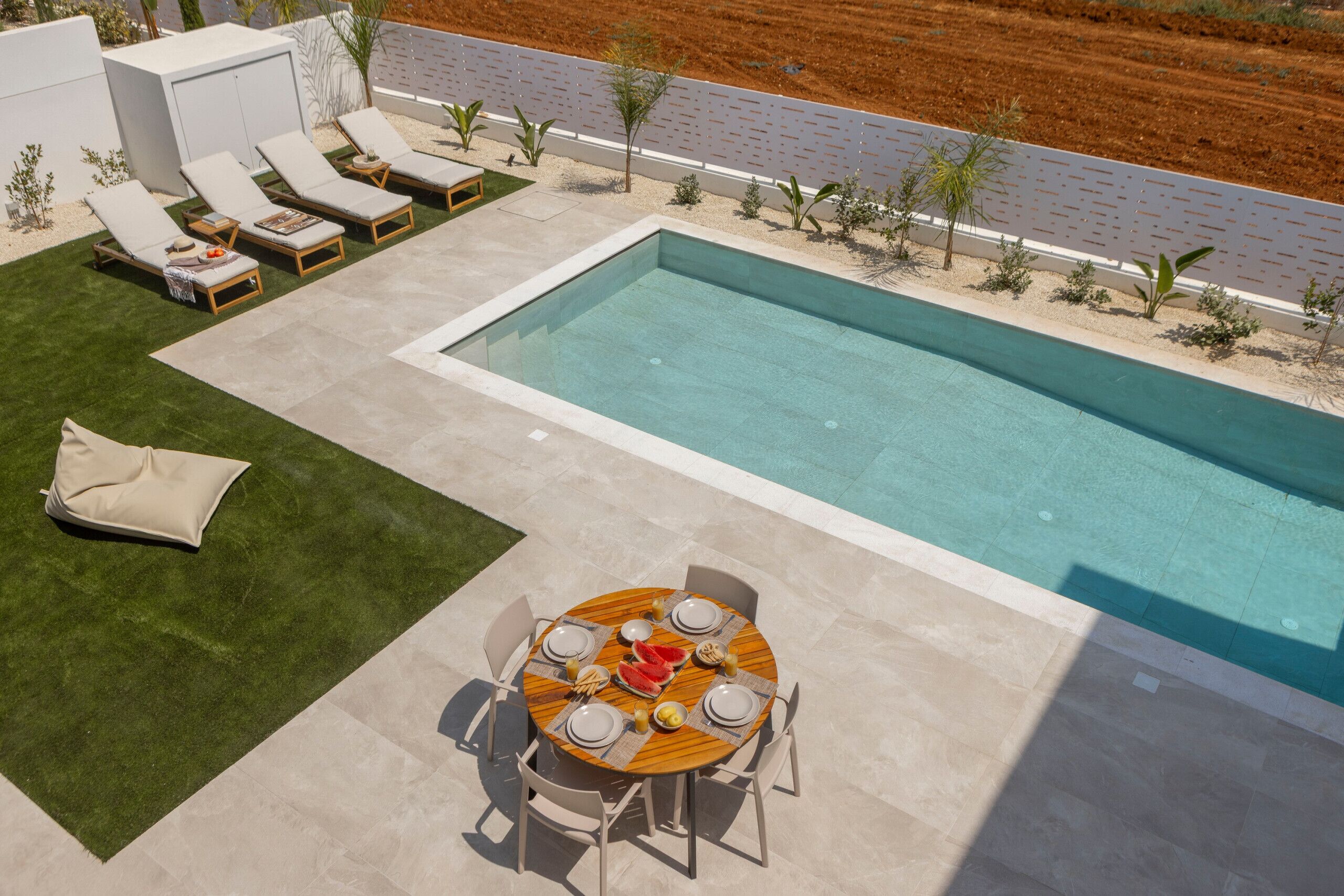Un espace piscine avec une table dressée pour quatre personnes, entouré de chaises longues et d’un petit jardin.