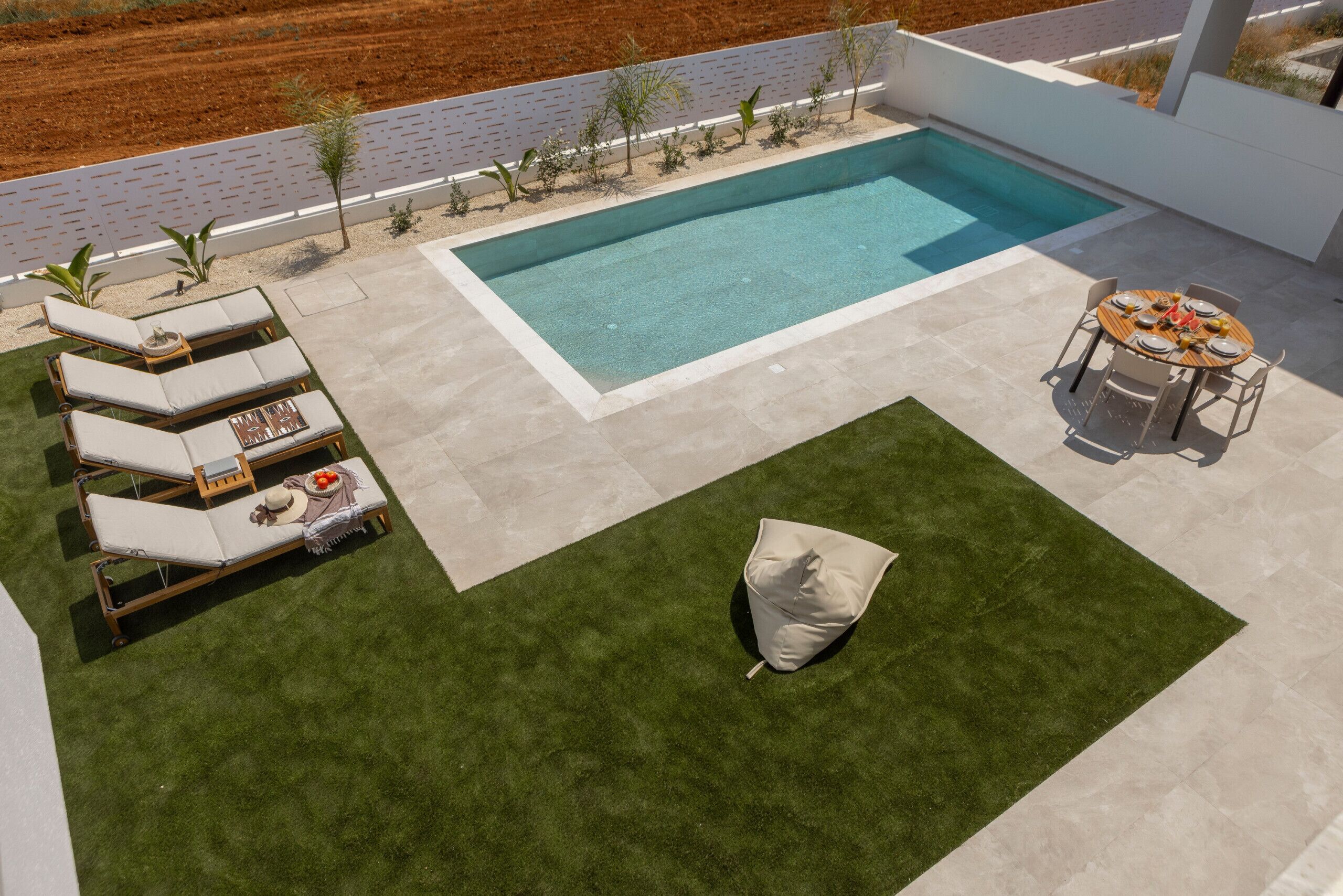 A modern outdoor pool area with a rectangular pool, lounge chairs, a dining table, and a small garden.