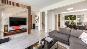 A modern living room with a flat-screen TV, a grey sofa, a coffee table, and a dining area with a table and chairs.