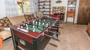 A room with a foosball table, a sofa, a chair, and a wooden shelf with decorative items.