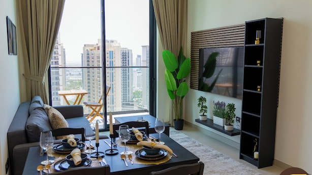 A modern living room with a dining area, large windows, a black entertainment unit, and a view of high-rise buildings. - Binghatti Gardenia High Floor Olympic  (Dubai)