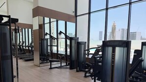 A gym with various exercise machines, large windows, and a view of a cityscape. - Binghatti Gardenia High Floor Olympic  (Dubai)