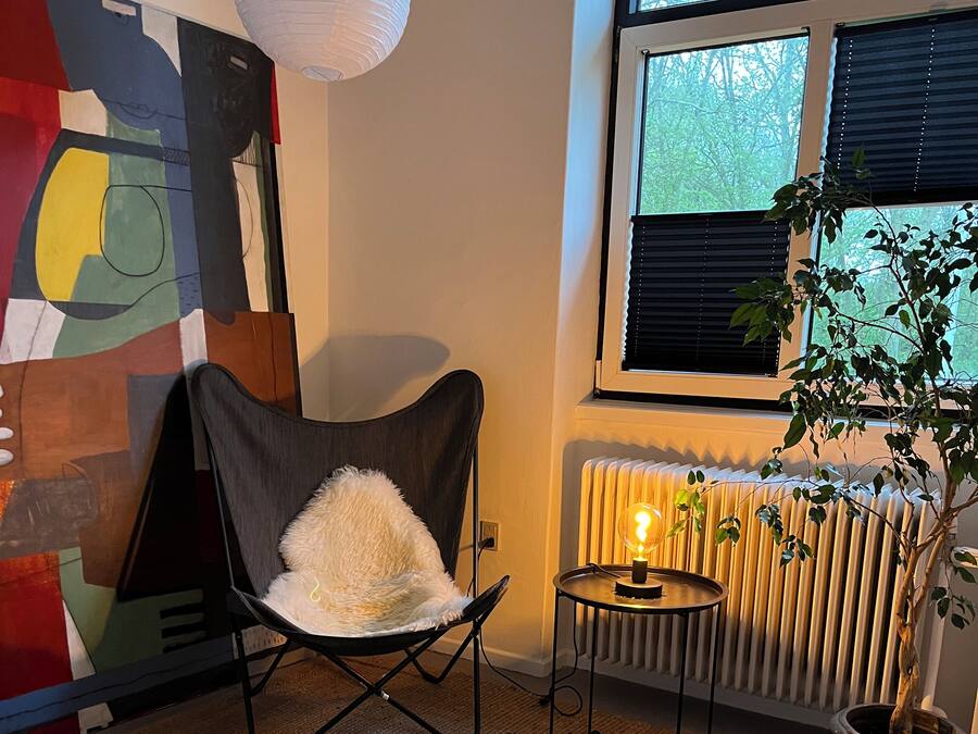 A room with a black chair, a small table, a radiator, a potted plant, and a large abstract painting on the wall.